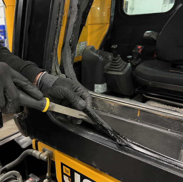 Technician removing old polyurethane sealant from JCB excavator cab frame using hand tools